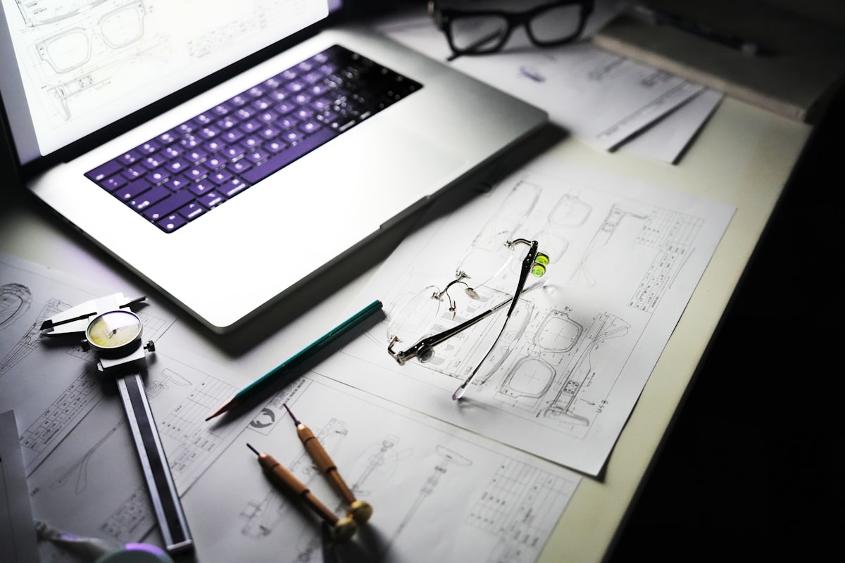 A design laid out on paper with dividers, a rule and a pencil beside a laptop